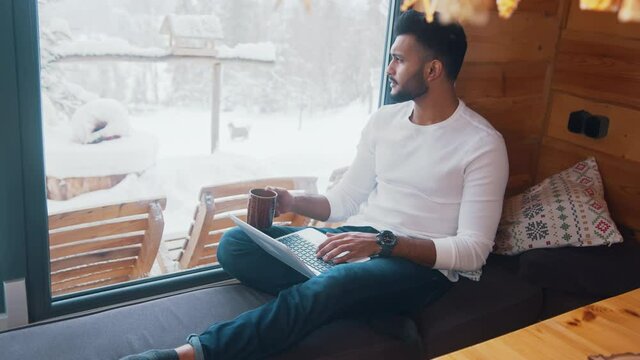 Young indian man relaxing near the window in cozy home, drinking coffee and using laptop. High quality 4k footage