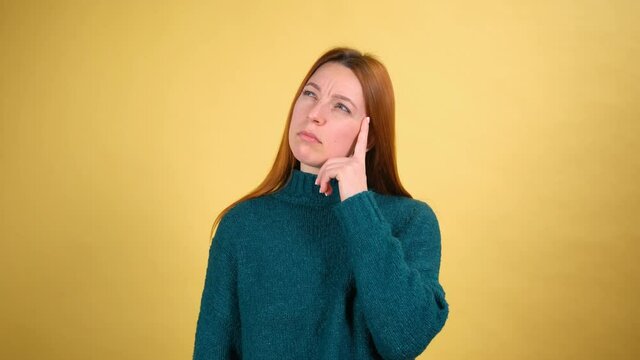Thoughtful young woman with red hair in green sweaters rubbing her chin and looking aside with pensive expression. pondering a solution, doubting question. Studio shot isolated on yellow background