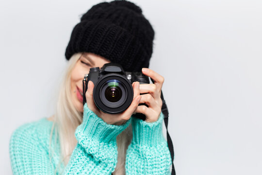Close-up portrait of pretty blonde girl taking photo on modern camera, wearing blue sweater and black hat on white background.