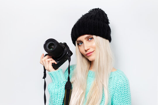 Studio portrait of young blonde girl holding photo camera, wearing blue sweater and black hat on white background.