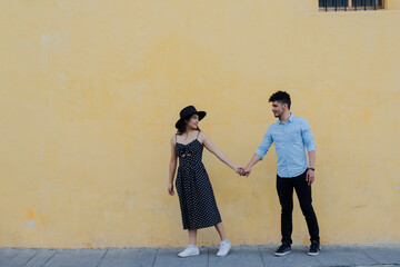 Couple walking hand in hand looking at each other on a big yellow wall - Hispanic couple on vacation walking in a colonial city - Tourists hand in hand on a yellow wall background