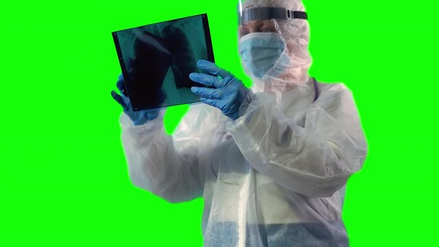 Doctor wearing a PPE suit, face shield and mask is inspecting a patient's X-ray of lungs as test on Covid with a phone using AR or taking a photo, on green background