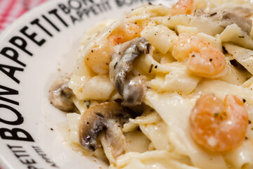 Delicious fettuccine with cream sauce of mushrooms and shrimp on a plate that says Bon Appetite with red checkered tablecloth.
