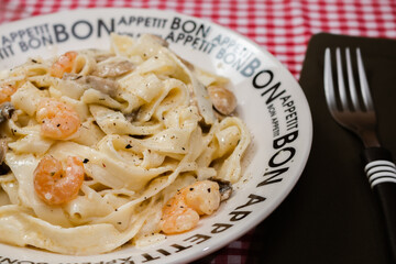 Delicious fettuccine with cream sauce of mushrooms and shrimp on a plate that says Bon Appetite with red checkered tablecloth.