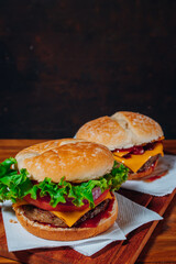 Delicious burgers with Bacon and cheddar cheese and with lettuce, tomato and red onion and bacon and cheddar on homemade bread with seeds and ketchup on a wooden surface and black background.