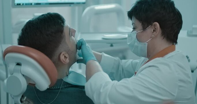 A male patient is sitting in the dentist chair, the female dentist is sitting next to him and exploring the patient&rsquo;s mouth cavity with her fingers.