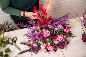 Fototapeta na wymiar Florist's workplace in a loft space. An experienced florist composes a bouquet of roses, carnations and eucalyptus, adding flowers step by step. She is tying a bouquet with a red ribbon. Tutorial