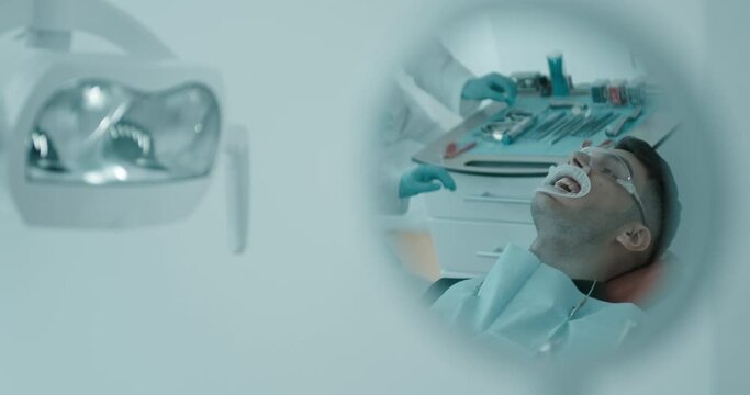 A mirror reflection of male Arabian patient who is having cheek retractor and protective eyewear on him. He is being examined during dentist`s appointment.