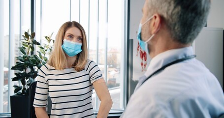 Fototapeta premium Rear of Caucasian senior experienced handsome professional male physician specialist sitting in clinic cabinet making injection to pretty female patient in medical mask vaccination coronavirus concept