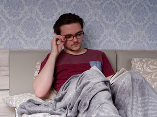 a young guy in a red T-shirt sits on a light-colored bed and reads a thick book