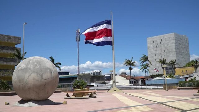Costa Rica flag with palm trees waved by the summer wind with the stone sphere and the legislative assembly in the background with the blue sky and clear clouds