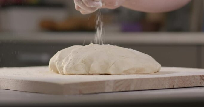 Cooking dough at home in the kitchen. The hand sprinkles flour on the dough for home baking of bread or pizza or cakes. Cinematographic 4K slow motion video.