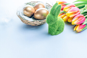 Egg color. Happy Easter decoration: golden colour eggs in basket with spring tulips, white feathers on pastel blue background. Foil minimalist egg design, modern template.