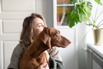 Woman owner and her Vizsla dog sitting together on windowsill, looking through the window. Love for...