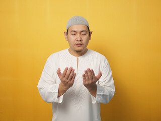 Portrait of Asian muslim man prays to God, praying gesture hands raised up, against yellow