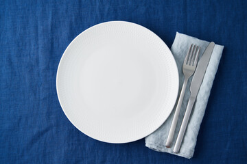 Clean empty white plate, fork and knife on bright blue dark linen tablecloth on table, copy space, mock up, top view. Concept for menu with utensil, blue textile napkin, fork and knife