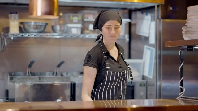 Middle shot of positive slim young woman in apron and cook hat putting plate with delicious vegan salad on counter and smiling looking at camera. Pretty chef in working cafeteria. Slow motion.