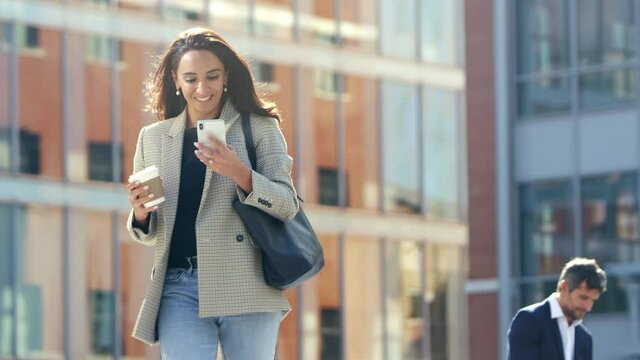 Smiling businesswoman with takeaway coffee walking to office following directions on mobile phone app - shot in slow motion