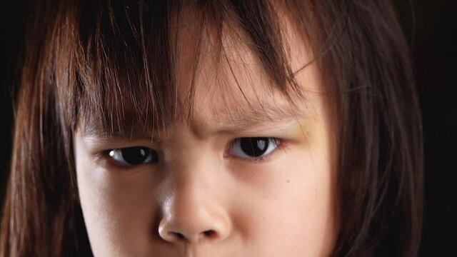 Close-up portrait of an angry cute little girl looking at camera with regret and displeasure in her eyes in the dark room at home. The concept of an unhappy childhood.
