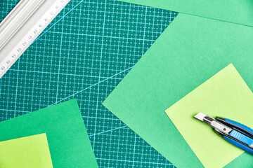 Green sheets of paper, a metal ruler and a cutter on a cutting mat