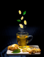 Glass of hot ginger tea with ginger slices (roots) and leaves placed on separate planks on a black background.