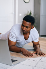 Fototapeta na wymiar Close-up view of young African-American man exercising workout in horizontal plank position using laptop to watching yoga and training video digital tutorial.