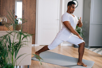 Focused fit African-American man making sport fitness exercise standing on yoga mat at bright domestic room, looking away. Concept of sport training at home gym.