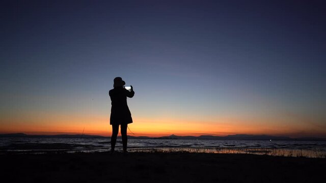 The silhouette of a woman standing with a phone and taking pictures of the lake before sunrise.