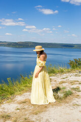 Fototapeta na wymiar A pretty girl in a long yellow dress standing on a high cliff by the bay or river on sunny summer day. Young beautiful woman with short hair and straw hat enjoying view on the shores of the big river