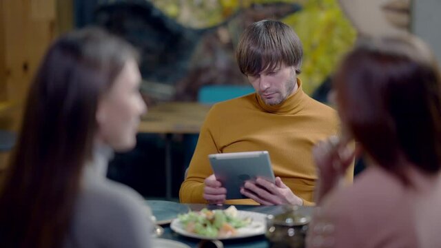 Absorbed confident Caucasian man surfing Internet on tablet as blurred women talking at front. Portrait of busy successful millennial dining with friends in restaurant. Slow motion.