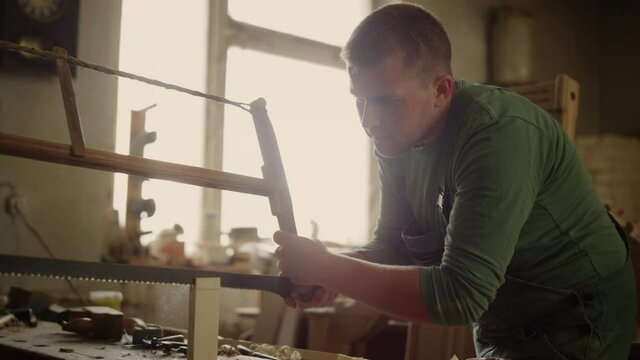 Focused man working with wooden plank indoors. Young man sawing wooden piece