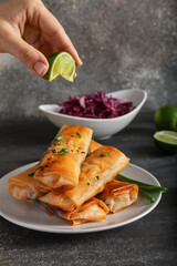 Woman squeezing lime into  plate with tasty fried spring rolls on dark background, closeup