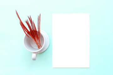 Square invitation card simulated with red grass flowers in a white glass with saucers on a light blue table.