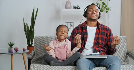 Funny caring afro american daughter little girl sitting with dad on sofa closes laptop of busy businessman wearing headphones pauses at work practices meditation lotus position relieves stress resting