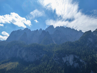 A panoramic view on high Alps in the region of Gosau, Austria. The lower mountains are overgrown with dense forest. The chains in the back are stony and barren. Few pine trees popping up. Sunny day.