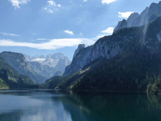Fototapeta na wymiar Panoramic view on Gosau lake, with Dachstein glacier in the back in Austrian Alps. The lake is surrounded by high mountains, overgrown with tall trees. Sun reflects on the surface. Serenity and calm