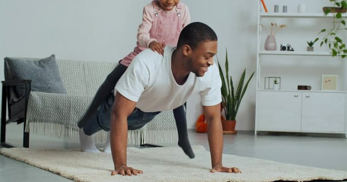 Afro-American single-parent family having fun at home, strong father doing sports during lockdown pushing up from floor on carpet while unexpectedly beloved daughter jumps on back of her daddy hugs