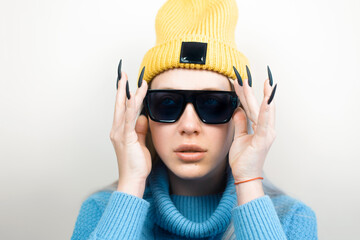 Young woman in black glasses, yellow hat and knitted sweater on light background. Teenage outfit