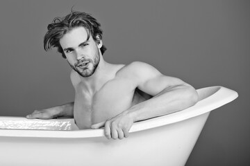 Man relax in bath, guy with muscular body and bare chest has fashion hair sitting in white bathtub,...