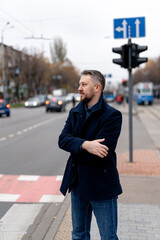 Thoughtful man in gray coat and dark suit with arms hugging elbows looks at the street. Autumn city road blurred background.