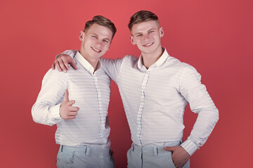 Brotherhood and friendship concept. Happy men hugging. Two brothers smiling and pointing finger. Models standing on red background. Twins wearing blue shirts and pants.