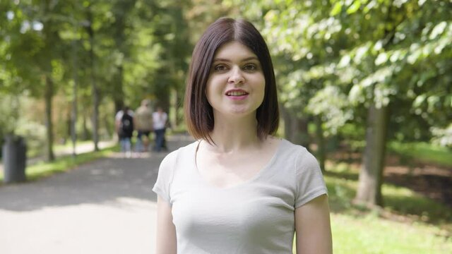 A young Caucasian woman talks to the camera on a pathway in a park on a sunny day