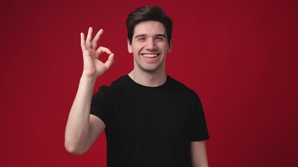 Funny surprised young man 20s years old wearing casual basic black t-shirt posing isolated on red color background in studio. People sincere emotions lifestyle concept. Say wow showing okay ok gesture