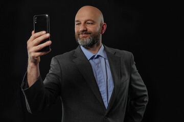 Portrait of a bald business man in a grey suit on a dark background. Model is in his 40s, with grey beard, blue shirt. Holding and looking at his phone. Video call concept.