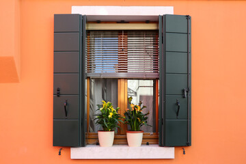 A picturesque window with open shutters on the bright orange wall of the house, pots with flowering calla lilies on the outside windowsill. Home style Italy