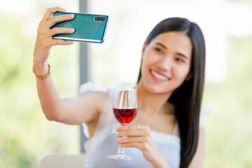 A beautiful young cute black long hair Asian woman holding red wine glass and use a smartphone to take selfie with happy smile face