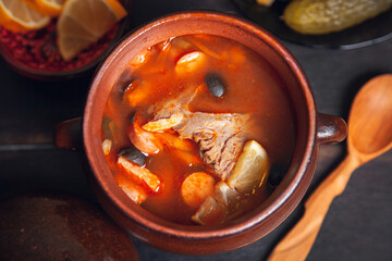 Homemade Traditional Russian soup Solyanka with meat, sausages, vegetables, pickles and olives with lemon in ceramic pot with wooden spoon on dark wooden background