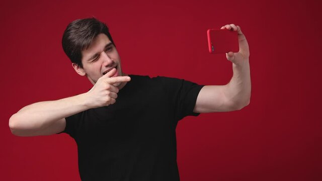 Funny young man 20s in basic black t-shirt posing isolated on red background studio. People lifestyle concept. Doing selfie shot on mobile phone point finger blinking showing thumb up biceps muscles