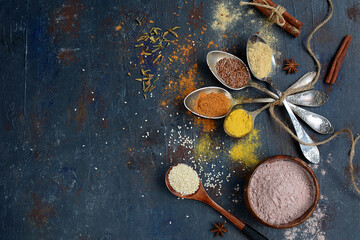 Fototapeta na wymiar Colorful spices, seeds and herbs for cooking and pastry in spoons and a bowl on a dark background with copy space. View from above.