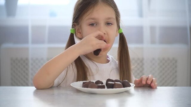 Little child girl sits at the table and eats chocolate candy.	
